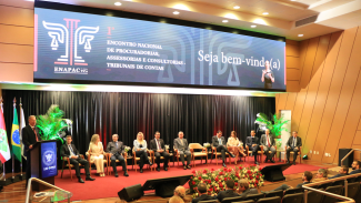 Foto horizontal que mostra o palco composto por autoridades durante o primeiro Enapac. Ao púlpito, fala o presidente Herneus De Nadal. Ao lado dele, outras 11 pessoas. No telão, ao fundo, identidade visual do evento.