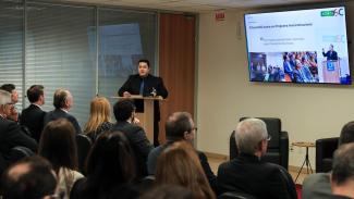Banner horizontal com foto do assessor da Presidência Flávio Alves falando ao microfone no púlpito do auditório. Ele é um homem branco, de cabelos castanhos. Veste terno e gravata. À sua frente, de costas, o público participante. Ao fundo, um televisor exibe a apresentação sobre o Acerta SC.