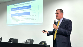Imagem mostra um homem de terno escuro, à direita, falando ao microfone. À esquerda há uma projeção na parede, com o título "Contexto Institucional Brasileiro"