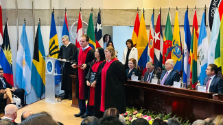 Foto mostra três pessoas em destaque no primeiro plano, mais ao centro da imagem. O fundo da foto é colorido, composto por diversas bandeiras colocadas em mastros