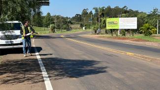 Imagem mostra uma rodovia. À margem esquerda, há um homem de colete verde com uma prancheta na mão. DO lado direito, há uma placa com branca e verde, com a expressão Santa Catarina em destaque, e com informações sobre a obra. O fundo se dá por árvores em uma parte e um céu azul.