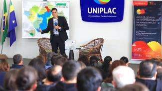 Imagem mostra um homem em um palco com um microfone à mão, com uma mapa ao fundo. À direta, há um cartaz em azul, alaranjado e amarelo.