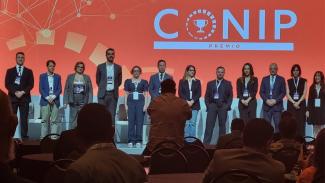 Foto horizontal com os representantes dos projetos finalistas do Prêmio Conip, ao fundo. Ao todo, há 12 pessoas, entre elas o auditor fiscal de controle externo Marcel Damato Belli, do TCE/SC (primeiro da esquerda para a direita), que estão em pé.  