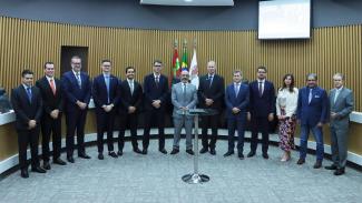 Banner horizontal com foto de autoridades reunidas no plenário do TCE/SC, com participantes da mesa de consensualismo e conselheiros alinhados lado a lado em frente a uma mesa central. Ao fundo, há painéis de madeira e bandeiras institucionais. No centro da imagem estão o defensor público-geral, Ronaldo Francisco, o secretário da Fazenda, Cleverson Siewert, o desembargador André Luiz Dacol e o presidente do TCE/SC, Herneus De Nadal, todos em trajes formais, posando para registro institucional.
