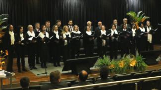 Foto de 22 participantes do Coral Hélio Teixeira da Rosa durante apresentação no palco do TCE/SC. Há homens e mulheres, um ao lado do outro, com pastas na mão. Todos vestem roupa preta. Como decoração, há alguns vasos de plantas.