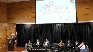 Foto de oito pessoas no palco do auditório do TCE/SC, durante a abertura do evento. O conselheiro Aderson Flores fala ao púlpito, em pé, à esquerda. À direita, sentados em poltronas, um ao lado do outro, estão Isabel Vasconcelos (TCE/S), Ítalo Augusto Mosimann (TRE/SC), Ayrton Teixeira (Ejesc), Patrícia de Moura (CGU), Ricardo Wagner Araújo (CGU), André Millioli (MPSC) e Paulo Salum (TCE/SC). No telão, ao fundo, projeção do logo do Seminário.