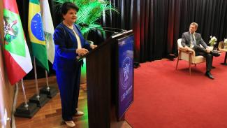 Foto horizontal ampla que mostra a palestrante Maria Sylvia Di Pietro falando ao público no púlpito do auditório do TCE/SC. Ela é uma mulher branca, de cabelos pretos e curtos. Veste roupa azul. Ao lado, em segundo plano, o conselheiro José Nei Ascari, mediador da palestra.