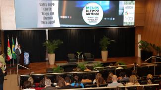 Banner horizontal com foto ampla do conselheiro José Nei Ascari discursando na abertura do evento, no palco do auditório do TCE/SC. Atrás dele, um telão que projeta o selo do desafio “Menos plástico no TCE/SC”. À frente dele, várias pessoas sentadas em poltronas, assistindo à sua fala. 