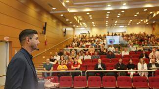 Banner horizontal com foto, de perfil e em primeiro plano, do auditor do TCU e palestrante do evento, Rafael Bittencourt. Ele é um homem branco, de cabelos e barba castanhos. Está em pé, no palco do auditório do TCE/SC, falando ao público. Em segundo plano, ao fundo, o público participante da palestra. Há várias pessoas sentadas em poltronas vermelhas do auditório.