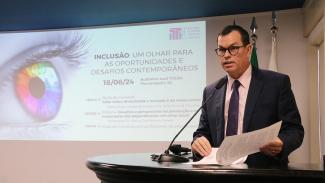 Banner horizontal com foto do conselheiro Adircélio de Moraes Ferreira Júnior durante a abertura do evento. Ele é um homem branco, de cabelos pretos. Veste terno e gravata e usa óculos de grau. Está em pé, diante do púlpito, falando ao microfone. Ao fundo, no telão, são projetados o logo e as informações do evento. 