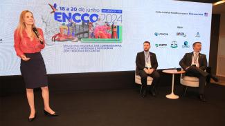 Foto da conselheira Cristiana Morais, do TCE/SP, que está de pé, com microfone de mão, palestrando, e dos servidores Rogério Guilherme e Geovane Cardoso, sentados em poltronas, ao lado, no palco do evento. A conselheira é loira de cabelos compridos e veste uma blusa vermelha de mangas compridas e saia preta. Os servidores do TCE/SC são claros, de cabelos castanhos e barba e estão de terno escuro. Ao fundo, telão com a logo do evento e das entidades apoiadoras.