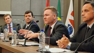 Banner horizontal com foto de quatro homens sentados diante da bancada de autoridades no evento. Entre os presentes, o conselheiro José Nei Ascari e o subprocurador-geral Paulo Locatelli. Ambos vestem terno e gravata.ntaçãoação