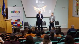 Foto do presidente Herneus De Nadal e do conselheiro da Uvesc Felipe Patzaff, no palco do auditório onde ocorre o evento. Ambos estão em pé. Herneus fala ao microfone, ao público, que está sentado à sua frente, de costas para a foto, em poltronas. No telão, projeção da apresentação com o texto “O Tribunal da Governança Pública”, e, ao lado, banner da Uvesc. 