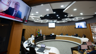 Banner horizontal com foto da bancada de autoridades no Plenário do TCE/SC. Na televisão, no canto superior esquerdo, a imagem do conselheiro Wilson Wan-Dall, participando de forma remota. 