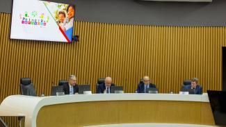 Foto do Plenário do TCE/SC, em que aparecem sentados em seus respectivos lugares, da esquerda para a direita, o conselheiro substituto Gerson dos Santos Sicca, e os conselheiros Luiz Eduardo Cherem, Wilson Wan-Dall, falando ao microfone, e José Nei Ascari. Todos vestem ternos azul-escuro. No fundo, no alto à esquerda, aparece uma tela de televisão fixada na parede, exibindo a logomarca do Painel Primeira Infância.