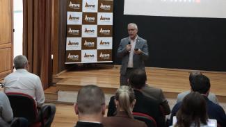 Foto horizontal do diretor Sidney Tavares falando ao público em pé, no palco do auditório. Ele é um homem branco, de cabelos grisalhos. Está vestindo paletó cinza. Ao fundo, banner com logo da Amve.