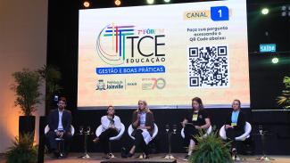 Imagem mostra cinco pessoas sentadas em poltronas brancas à frente de um telão que mostra o logotipo do Fórum TCE Educação, com predominância do tom amarelo. Há um homem, à esquerda, e quatro mulheres na imagem
