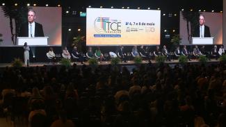 Foro mostra um palco com 14 pessoas sentadas uma ao lado da outra, de frente para a plateia. Ao fundo há três telões. O do centro mostra a logomarca do TCE Educação e do evento. Os laterais mostram um homem falando ao microfone.