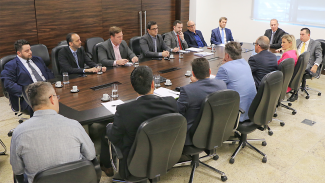 Banner com a foto dos 15 participantes — 14 homens e 1 mulher — da reunião. Eles estão sentados em volta de uma mesa. No canto direito da imagem, estão quatro conselheiros do TCE/SC. 