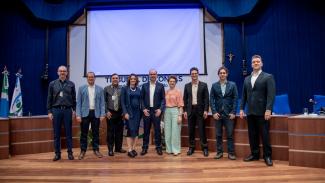 Foto do auditório do TCE/MS, onde aparecem nove pessoas, de pé, enfileiradas, entre elas, o diretor Nilsom Zanatto e os demais técnicos dos dois tribunais. Ao fundo, um telão sem imagens, em branco. E no canto esquerdo as bandeiras do Estado do Mato Grosso do Sul e do TCE/MS. 