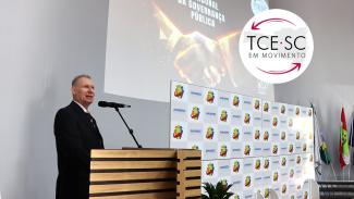 Imagem mostra o presidente do TCE à esquerda, vestindo um traje preto. Ao fundo, há uma projeção com a inscrição "TCE/SC, parceiro do bom gestor".