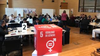 Imagem mostra um grupo de pessoas trabalhando em uma sala grande, em mesas numeradas. Em primeiro plano, há um cubo vermelho com o número 5 em branco. Cada uma das faces, além do numeral, tem a inscrição em espanhol Igualdad de Género e os símbolos que representam os sexos feminino e masculino com um sinal de igual na intersecção de ambos.