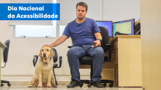 Foto de Guido, sentado, a frente da mesa, em seu ambiente de trabalho no TCE/SC. Ao seu lado, está a cão-guia, Coral. No canto superior esquerdo há uma tarja azul com o título "Dia Nacional de Acessibilidade".