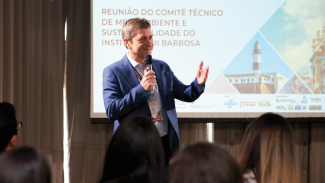 Foto do conselheiro José Nei Alberton Ascari fazendo apresentação durante a reunião do Comitê Técnico de Meio Ambiente e Sustentabilidade do Instituto Rui Barbosa. Ele está em pé, com terno azul. A sua frente, pessoas sentadas. Atrás, um painel com identificação da reunião. 