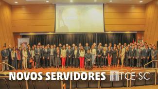 Os 61 novos servidores (mulheres e homens), em pé, e um cadeirante, no palco do auditório do TCE/SC. Ao fundo e no alto, telão, no lado esquerdo, banner com o logotipo do TCE/SC sobre fundo branco. A frente, cadeiras vazias.