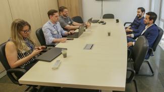 Imagem mostra cinco pessoas sentadas à mesa. À esquerda estão três pessoas com computadores. A primeira é uma mulher loura. Os outros dois são homens. À direita dois homens.