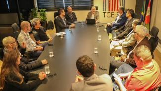 Foto horizontal com os participantes da reunião, entre representantes do TCE/SC, da SCTI, do Ciasc e da Udesc. Ao todo, há 13 pessoas — sendo uma mulher —, que estão sentadas ao redor de uma mesa. Na mesa, há copos com água, óculos, notebooks, entre outros objetos.
