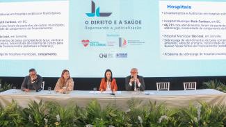 Imagem mostra um painel azul ao fundo com projeções e o nome do evento, "O Direito e a Saúde". À frente, uma mesa com quatro pessoas, dois homens e duas mulheres. 