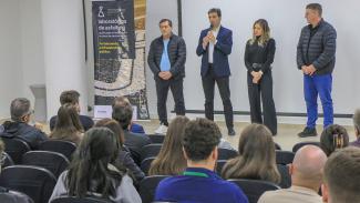Banner horizontal com foto de comitiva do TCE/SC no palco de um auditório. Há quatro pessoas, lado a lado. Entre elas, o diretor Rogério Loch fala ao microfone. Ao lado dele, banner do evento, com o título "Laboratórios de asfalto". À sua frente, o público participante, sentado em poltronas.