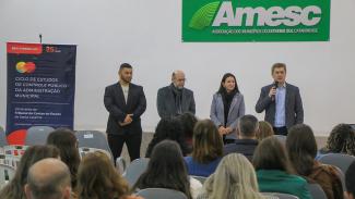 Imagem mostra público em primeiro plano. Ao fundo, com uma parede branca, há um palco com quatro pessoas. À esquerda há um cartaz em azul escuro e laranja e, no alto, mais para a direita, um cartaz escrito Amesc.