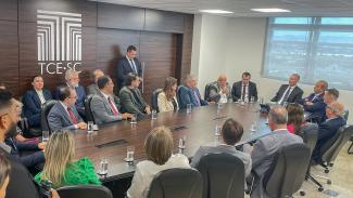 Foto das várias autoridades sentadas diante de uma mesa de reuniões enquanto conversam. Entre os presentes, os conselheiros Herneus De Nadal e Luiz Eduardo Cherem e o governador Jorginho Mello. 