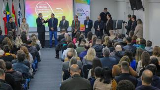 Imagem mostra em primeiro plano pessoas de costas sentadas em um auditório e olhando para um palco. Ao fundo, há 10 pessoas em pé olhando para essa plateia. No centro, o presidente do TCE fala ao microfone.  