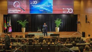 Banner horizontal com foto do especialista em Gestão Pública Fabrício Colombo durante palestra no auditório do TCE/SC. Ele está em pé ao fundo, no palco, falando ao microfone. À sua frente, de costas para a foto, a plateia participante. Ao fundo, no telão, é exibido o banner da palestra, com o título "Implementação de Corregedorias Municipais".