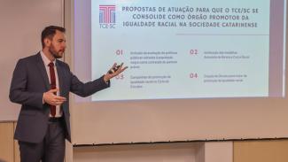 Foto do servidor Geovane Eziel Cardoso defendendo sua dissertação de mestrado. Ele é um homem branco, de cabelos e barba castanhos. Veste terno e gravata e está em pé. Atrás dele, projeção da apresentação.