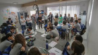 Um grupo de pessoas aparece em pé, ao fundo, em uma sala de aula. Eles estão de frente para um grupo de crianças.
