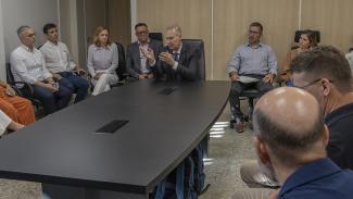 Foto horizontal que mostra uma reunião institucional em sala do TCE/SC, com participantes sentados ao redor de uma mesa retangular. Ao centro, o presidente Herneus De Nadal, em destaque, gesticulando enquanto fala. Ao redor, os demais acompanham a reunião com atenção. Na foto aparecem os diretores Sidney Tavares Junior, Rogério Loch e Marina Ferraz, além da chefe de gabinete Juliana Francisconi. 
