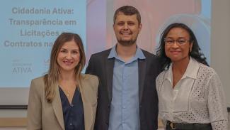 Foto horizontal que mostra os servidores do TCE/SC Marina de Miranda, Gabriel Schiochet e Elusa Silveira posicionados lado a lado, sorrindo para a câmera, em ambiente interno de evento. Ao fundo, há uma tela de apresentação com o tema “Cidadania Ativa”, compondo o contexto institucional da atividade. 