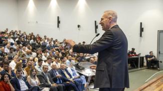 Imagem mostra em primeiro plano um homem falando ao microfone e vestindo um paletó escuro. Ao fundo, há uma plateia olhando para ele