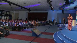 Foto da coordenadora do Lince, auditora fiscal de controle externo Tatiana Custódio, palestrando no nono Encontro Nacional dos Tribunais de Contas. O palco é uma estrutura em tons de azul e roxo. Ela está em pé, falando ao microfone.  Ela para uma palestra formada por homens e mulheres.