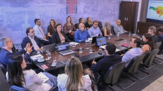 Banner horizontal com imagem de reunião institucional em sala do Tribunal de Contas de Santa Catarina. Ao centro, uma mesa retangular reúne diversos participantes, com notebooks e documentos. À esquerda, o conselheiro substituto Gerson Sicca conduz a reunião. Ao fundo, há a identidade visual do TCE/SC na parede e, à direita, uma tela exibe um mapa com dados. Os demais participantes, integrantes de entidades ligadas à educação, acompanham a reunião