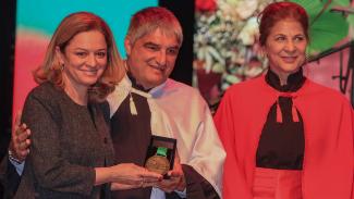 Foto horizontal que mostra a chefe de Gabinete da Presidência do TCE/SC, Juliana Cardoso, recebendo a medalha pelas mãos do reitor da Udesc, José Fernando Fragalli, e da vice-reitora, Clerilei Aparecida Bier. Eles estão um ao lado do outro, e sorriem. 