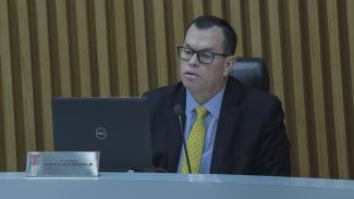 Banner horizontal com a foto do corregedor-geral do TCE/SC, conselheiro Adircélio de Moraes Ferreira Júnior, durante sessão plenária no TCE/SC, sentado à bancada, utilizando microfone e falando enquanto olha para a tela de um notebook. Ele é um homem branco, de cabelos pretos. Veste terno e gravata, usa óculos de grau e está em ambiente institucional com painel de madeira ao fundo. 