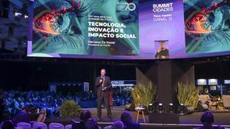 Banner horizontal com foto do presidente do TCE/SC, conselheiro Herneus De Nadal, durante palestra no Summit Cidades, realizado no Centrosul, em Florianópolis. O conselheiro aparece no palco, segurando um microfone e falando ao público. Ao fundo, grandes telões exibem a identidade visual da apresentação, com o tema “Tecnologia, Inovação e Impacto Social”, além de imagens gráficas e informações do evento. A plateia acompanha a exposição em um ambiente iluminado por luzes cênicas.