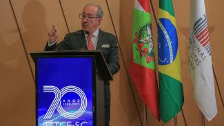 Foto horizontal do palestrante José Juis Garcia Guerrero que está falando do púlpito. Ele é um homem branco, usa óculos e veste um terno cinza escuro. Atrás dele, as bandeiras de Santa Catarina, do Brasil e do TCE/SC.