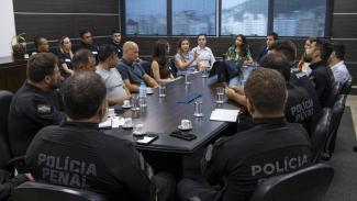 Imagem mostra várias pessoas ao redor de uma mesa. De frente ao centro, há quatro mulheres. De costas há homens com roupas pretas que nas costas aparece escrito Polícia Penal