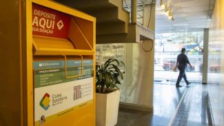 Imagem mostra, em primeiro plano, um recipiente amarelo, à esquerda. Ao fundo, à direita, há uma porta de vidro por onde está passando uma pessoa